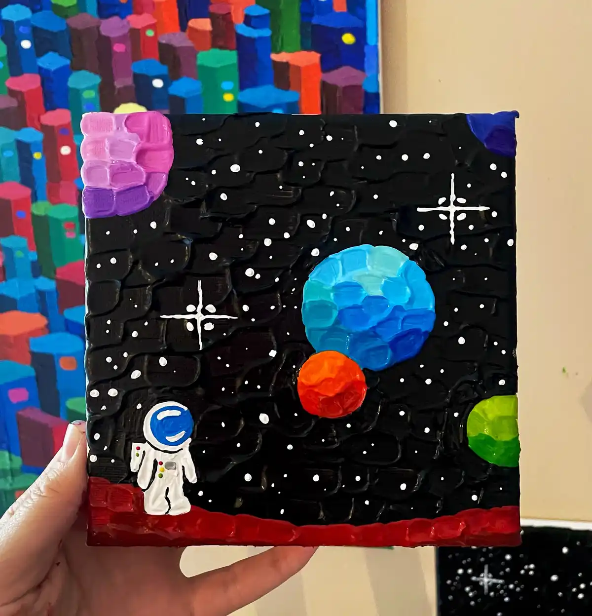 A hand holds a small, textured painting depicting a space scene with a white astronaut on a red planet, surrounded by…