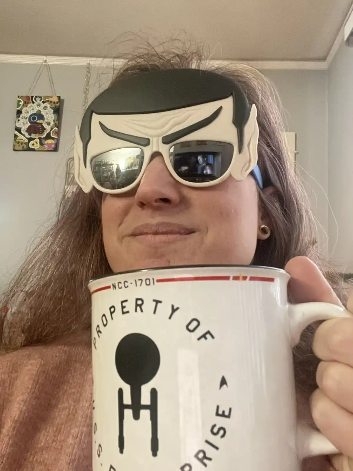 A person with long brown hair wears a large, white, stylized Spock mask with sunglasses and holds a white mug that reads…