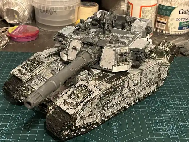 A heavily detailed, unpainted grey plastic tank model sits on a green cutting mat, covered in white paint splatters…
