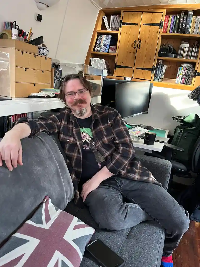 A man with glasses and a mustache sits relaxed on a grey couch in a cluttered home office, surrounded by bookshelves, a…
