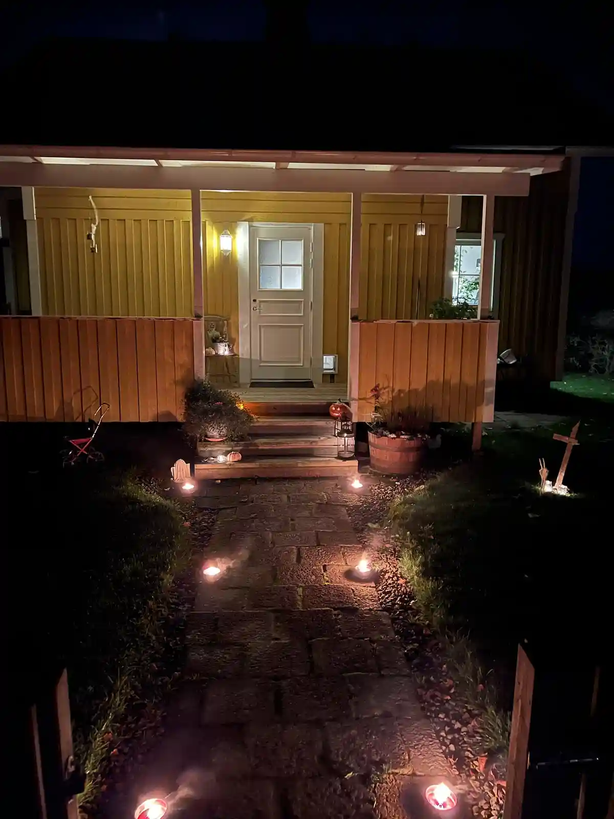 A nighttime photo of a house's front porch decorated for Halloween, featuring lit candles along a stone pathway, pumpkins…