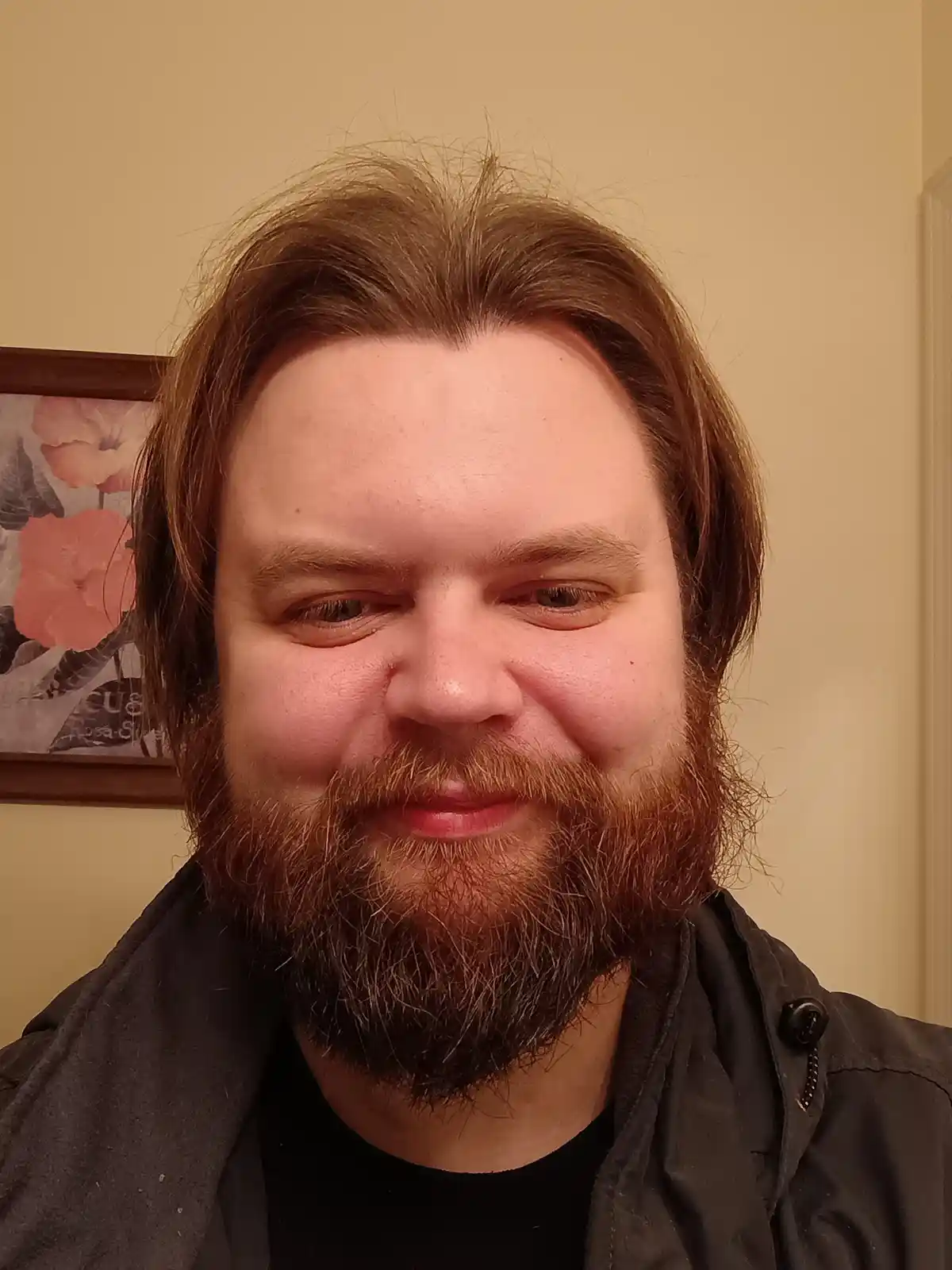 A close-up selfie of a man with reddish-brown hair and a full beard, wearing a dark jacket over a black shirt, looking…