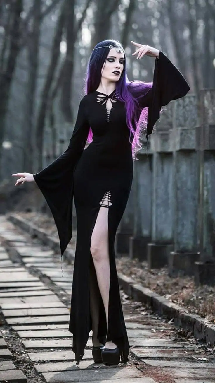 A woman with long purple hair and dark makeup stands on a stone path in a forest, wearing a black gothic dress with high…