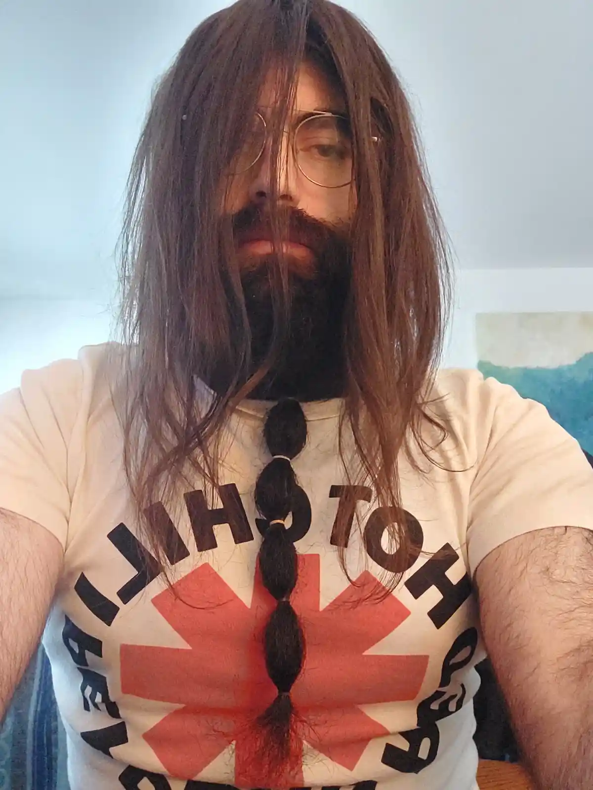 A person with long brown hair and a beard wears glasses and a white t-shirt with a red cross and black text. Their hair is…