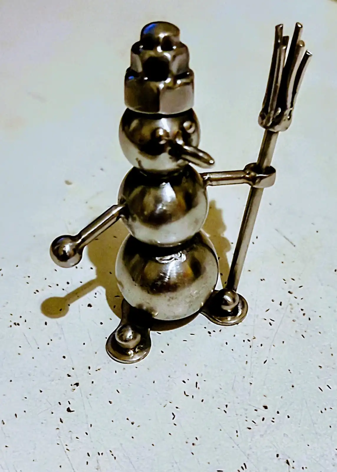 A metallic snowman figurine crafted from various kitchen utensils, including a nut for a hat, a spoon for a nose, and a fork…