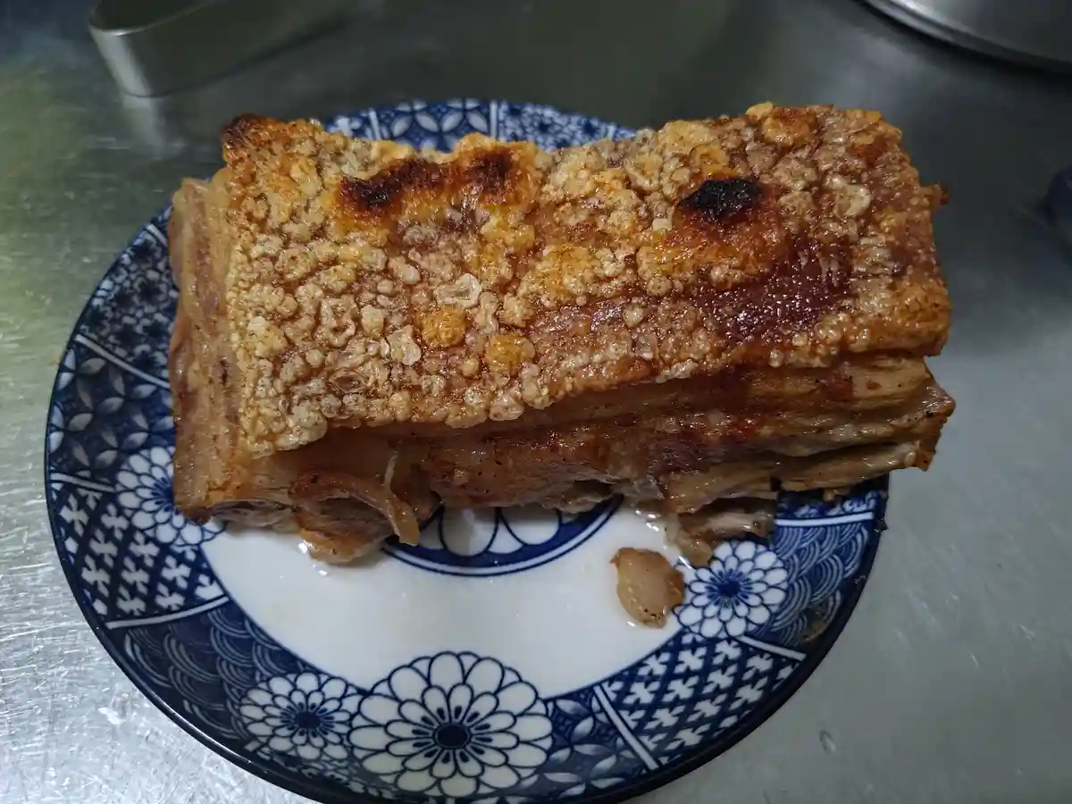 A close-up photo shows a thick, crispy-skinned slab of cooked pork belly resting on a blue and white patterned ceramic…