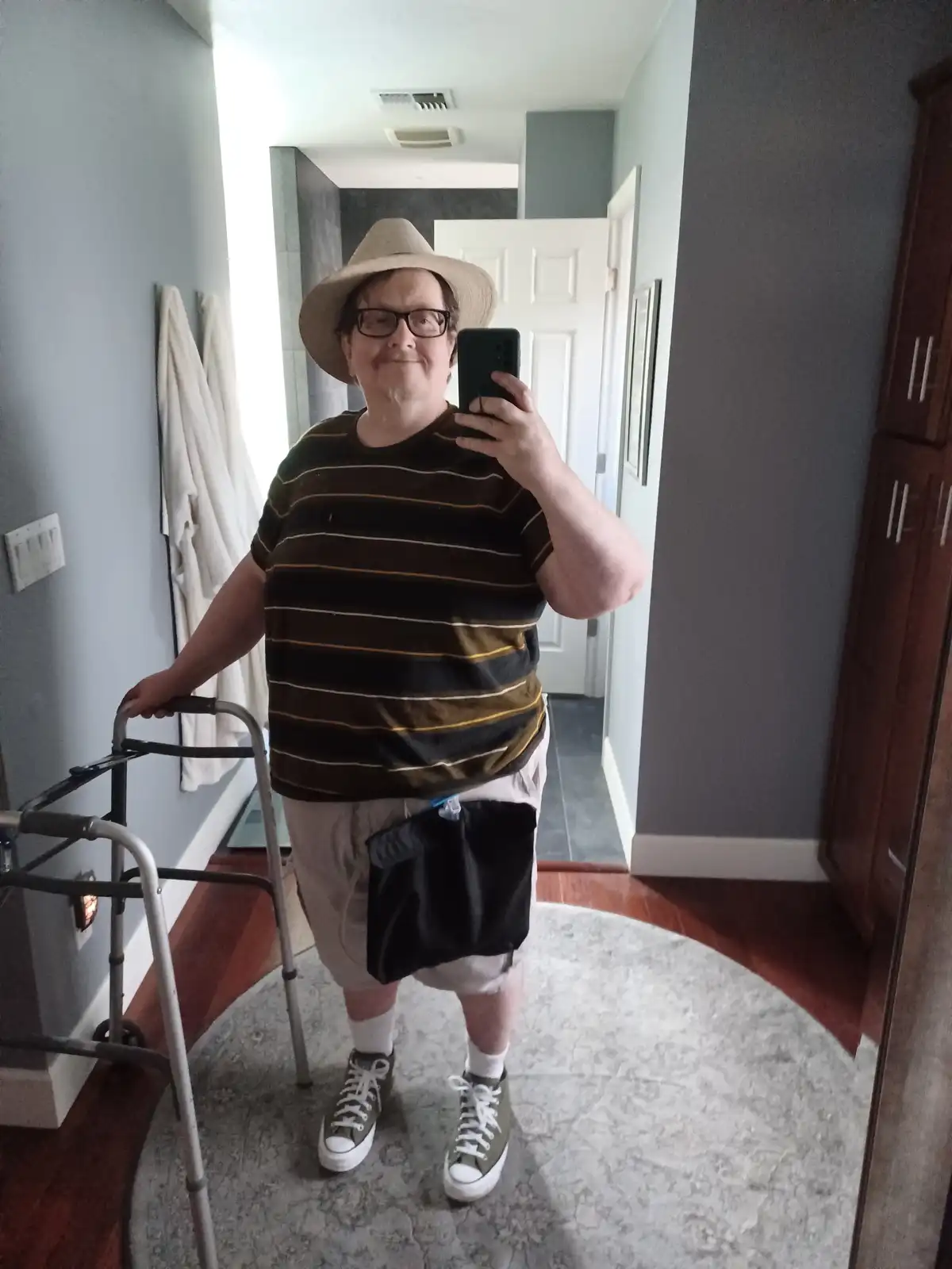 A person wearing a straw hat, glasses, a striped shirt, shorts, and sneakers stands in a hallway holding a walker while…