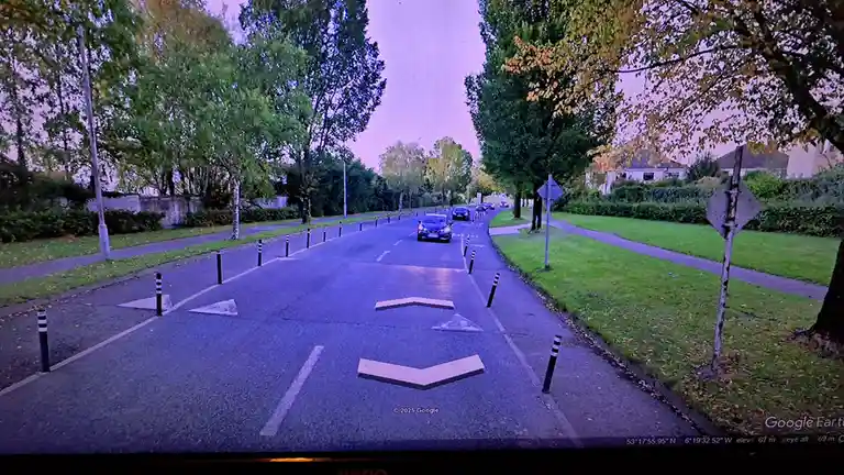 A Google Earth Street View image shows a paved road lined with trees and black-and-white striped bollards, with cars visible…