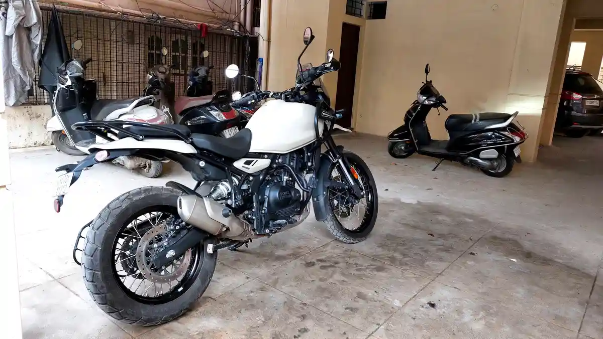 A white Royal Enfield motorcycle is parked in a covered garage alongside several scooters and a car. The overlaid text reads…