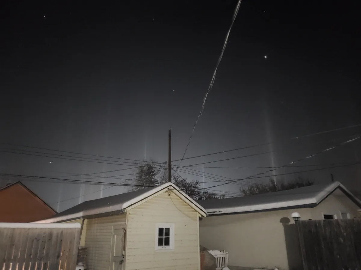 A nighttime photo of suburban houses with snow on roofs, power lines, and a dark sky with faint vertical light streaks…
