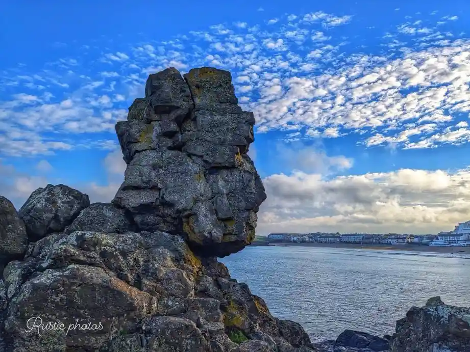 A scenic photo of a large, jagged rock formation by the seaside under a bright blue sky with scattered clouds. The rock has…