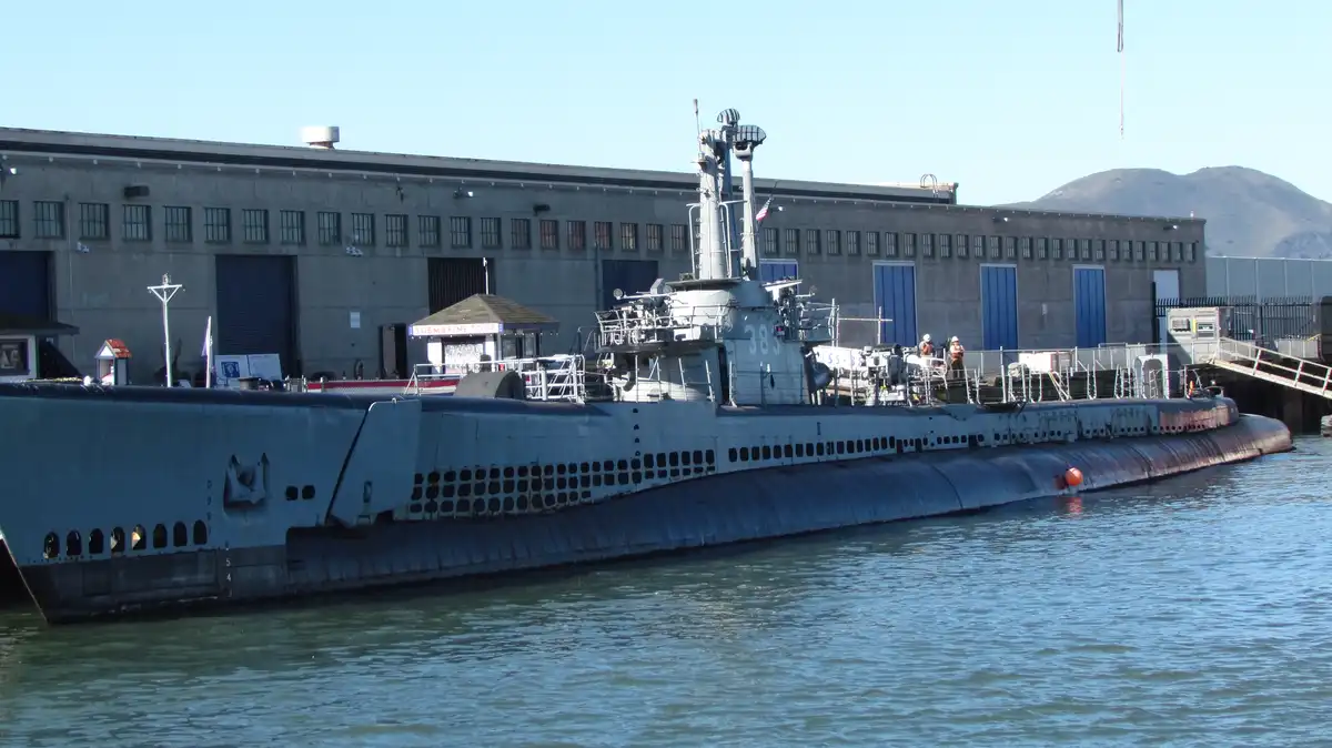 A gray submarine, marked with the number 383 and labeled 'SS-383', is docked at a pier next to a large industrial building…