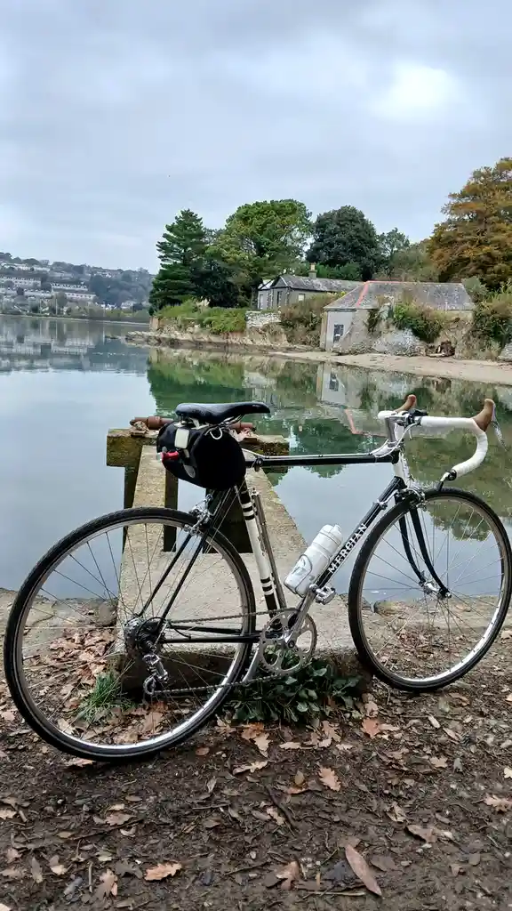 A black and white road bicycle with a water bottle and saddlebag is leaning against a stone pier beside a calm river. Across…