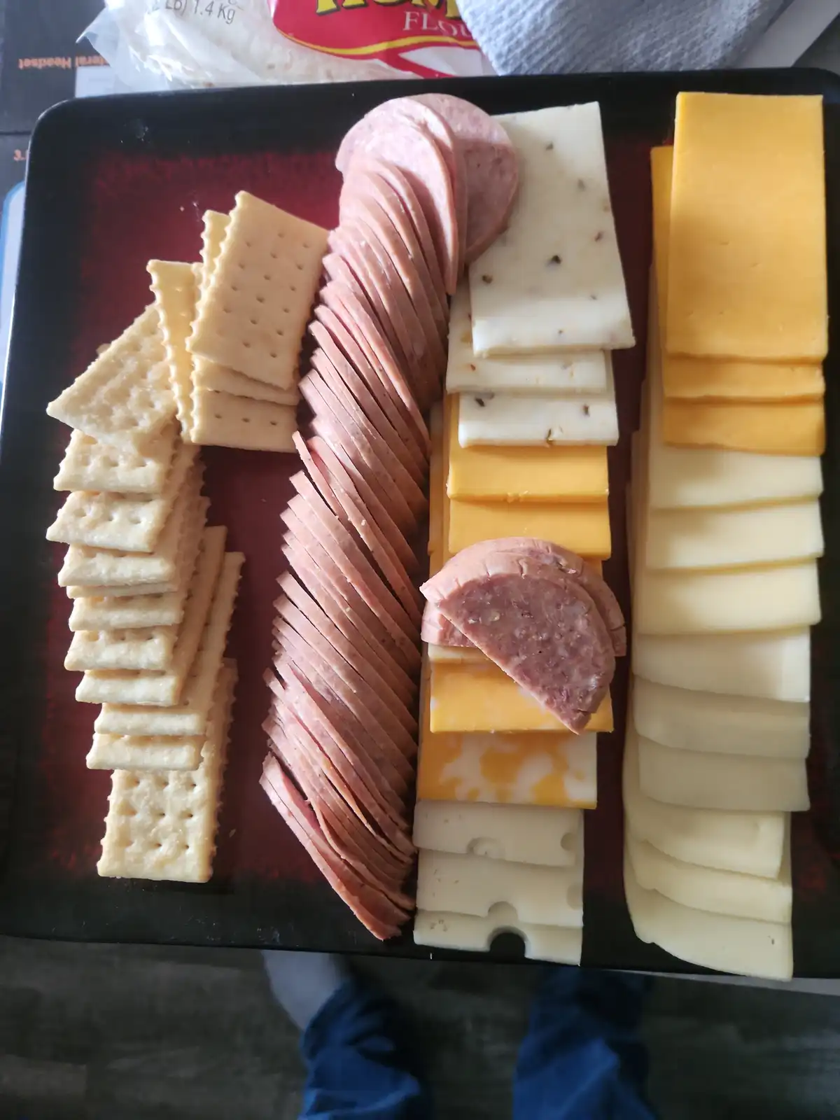 An overhead photo of a black tray holding a charcuterie board with neatly arranged crackers, sliced deli meat, and various…