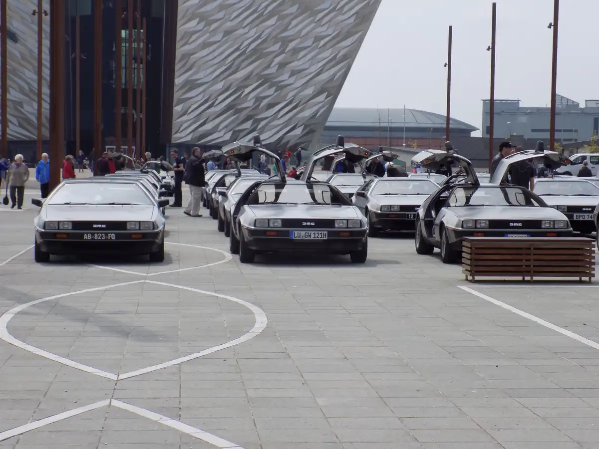 A row of silver DeLorean cars with gull-wing doors open, parked in front of a modern building with a geometric facade…