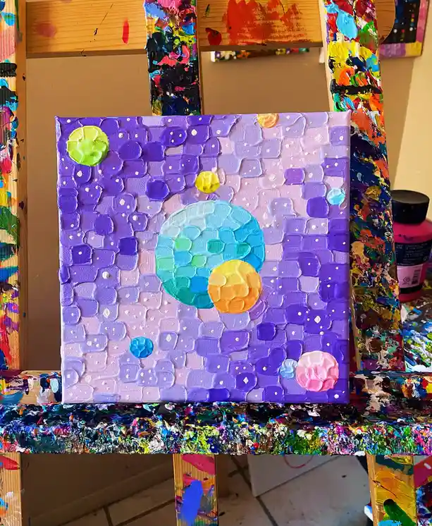 A textured, thickly painted canvas on an easel depicts a colorful, abstract solar system with several planets in pastel and…