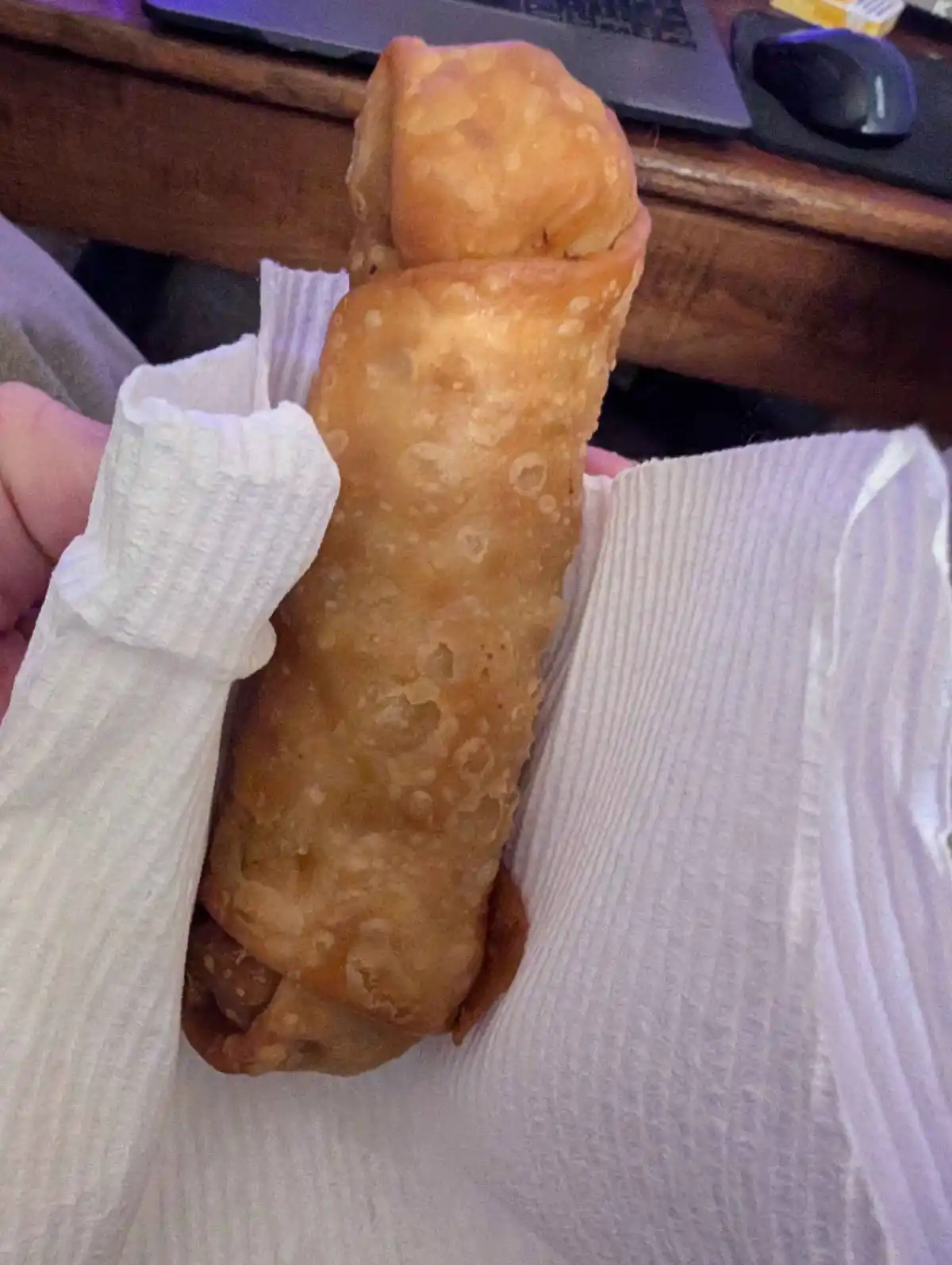 A hand holds a very large, golden-brown fried egg roll wrapped in white paper towels, with a laptop and mouse visible on a…