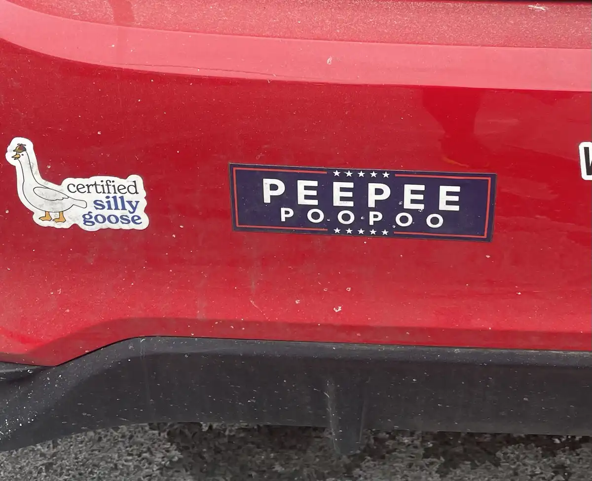 A close-up of a red car's rear bumper shows two stickers: one is a cartoon goose with the text 'certified silly goose', and…