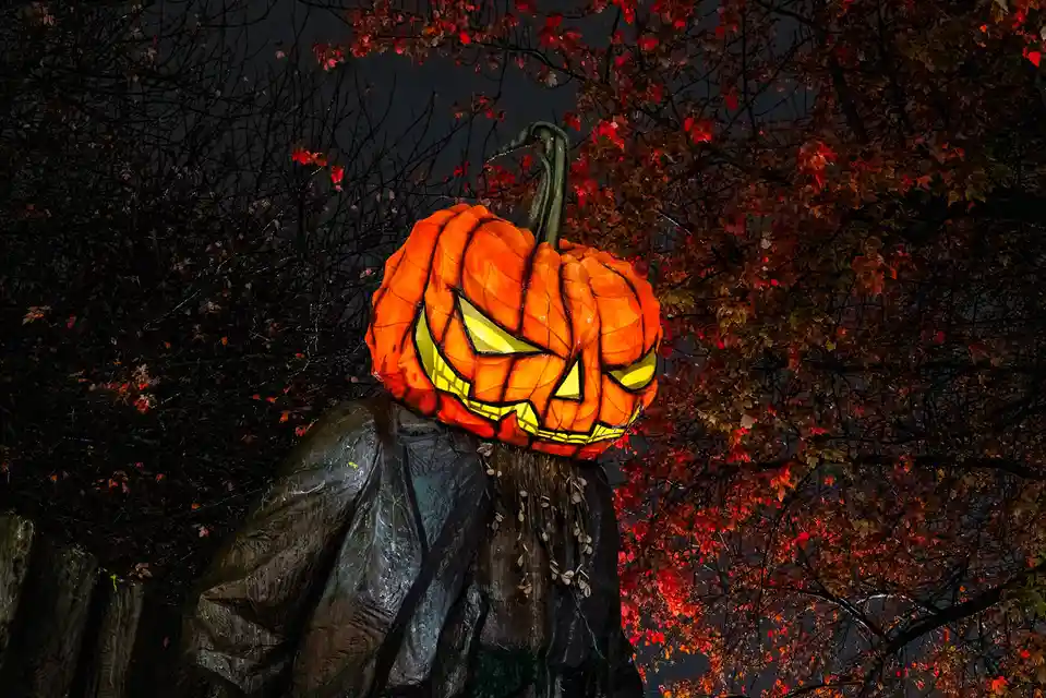 A large, illuminated jack-o'-lantern with glowing yellow eyes and a jagged grin is mounted on a dark, hooded figure…