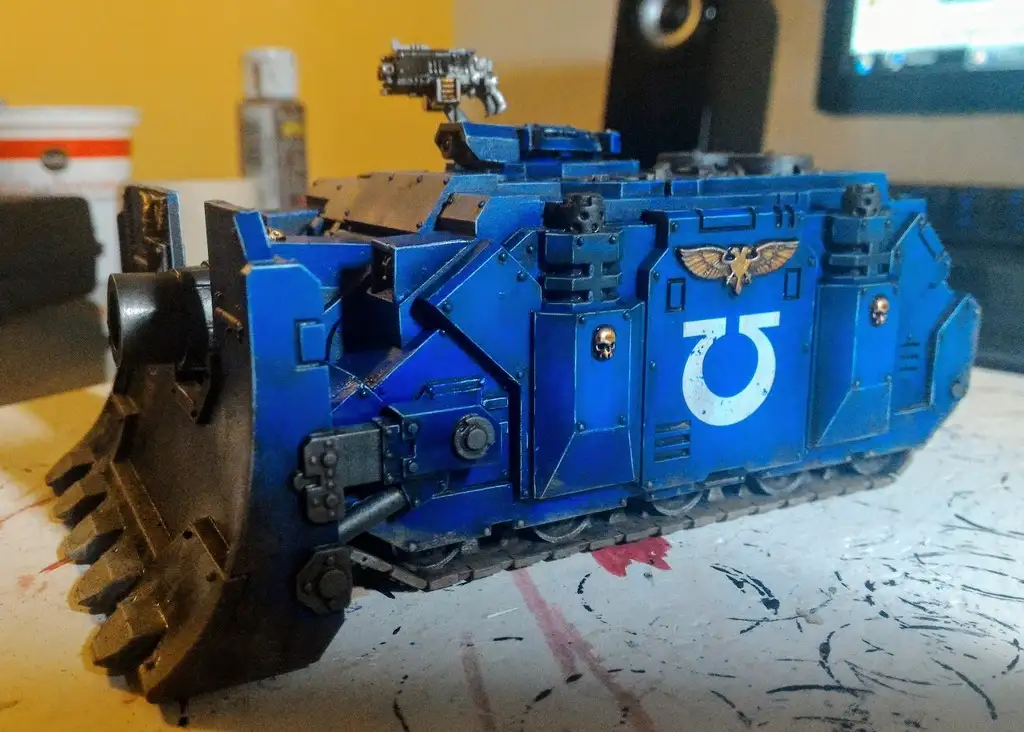 A blue-painted, heavily detailed miniature tank model with a large front scoop, mounted weapons, and a white symbol on its…