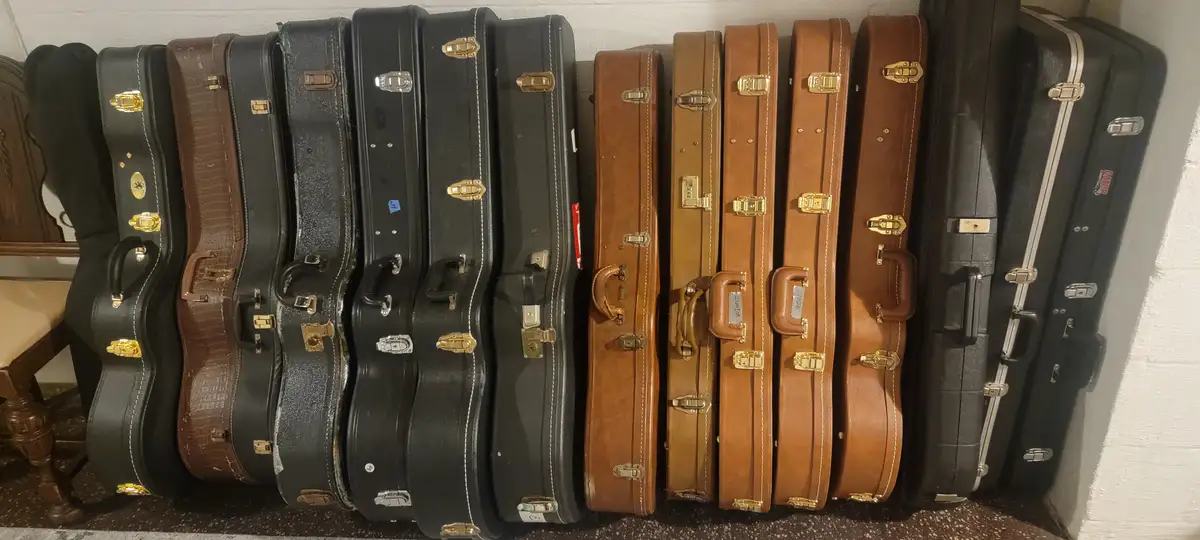 A row of guitar cases, mostly black and brown, are lined up against a wall on a speckled floor. The cases vary in style and…