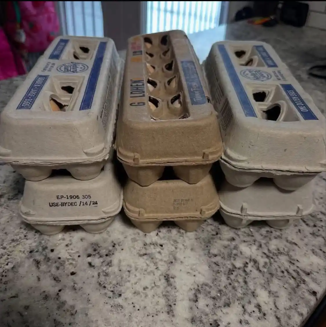 Three empty cardboard egg cartons sit side by side on a speckled countertop. The carton on the left has a visible 'USE-BY…