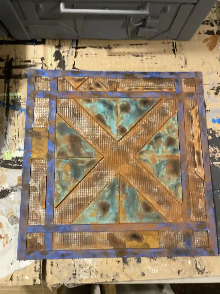 A square metal panel with a geometric, rusted, and weathered design featuring a central cross pattern of triangles and a…