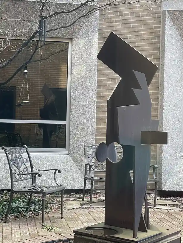 An outdoor photo shows a dark, angular abstract metal sculpture standing on a pedestal in front of a brick and stucco…