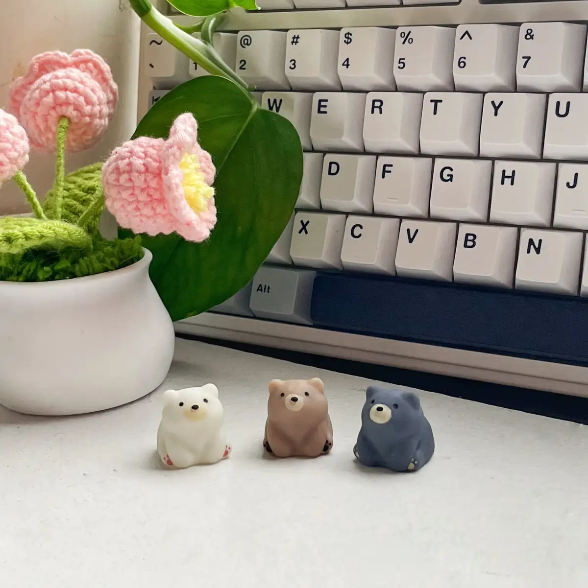 Three small, cute bear figurines in white, beige, and blue sit in front of a white keyboard on a desk, next to a crocheted…