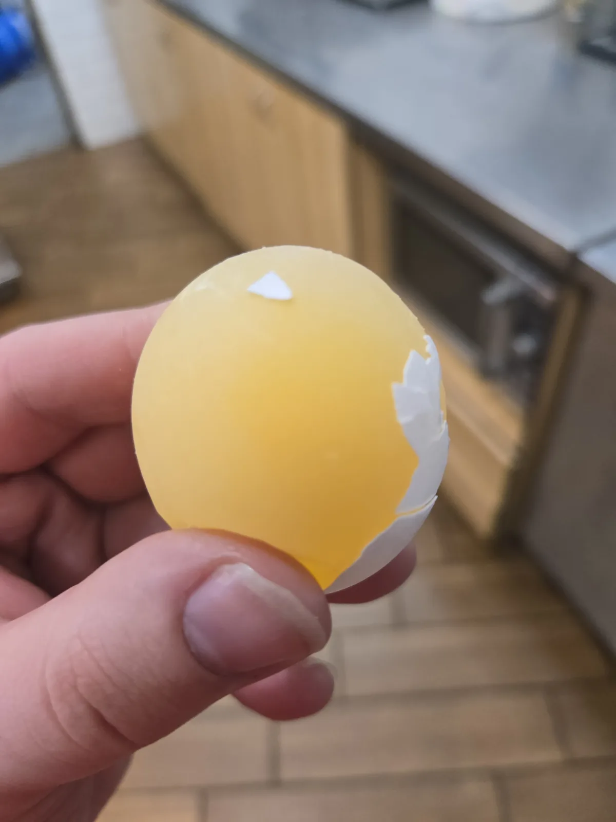 A hand holds a partially peeled hard-boiled egg, revealing mostly yolk with a small white shell fragment on top and a larger…