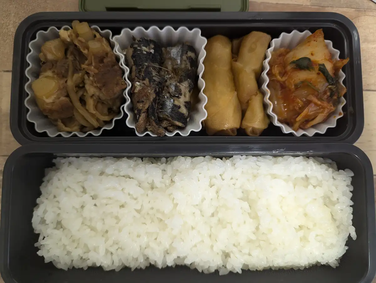 An overhead shot of a black bento box with white rice in the bottom compartment and four compartments of side dishes in the…