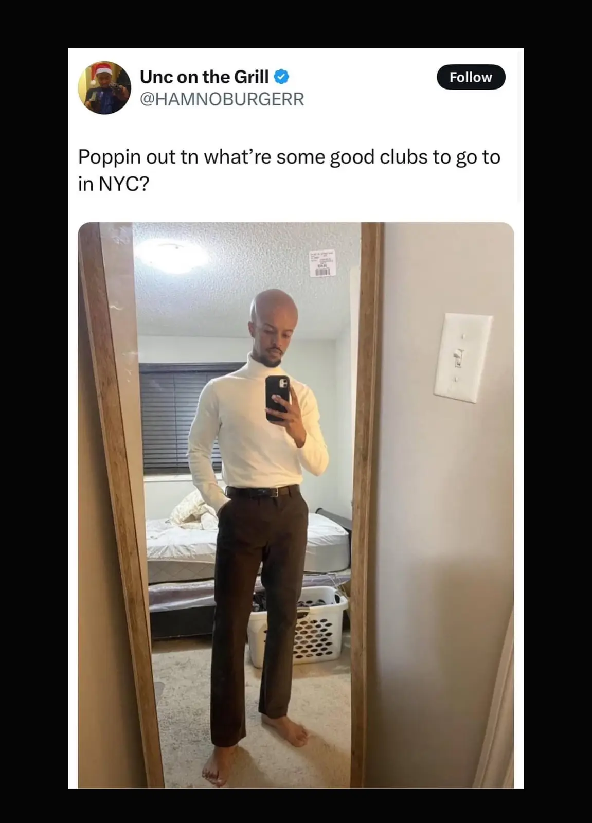 A man takes a mirror selfie in a bedroom wearing a white turtleneck and dark pants, looking stylishly prepared for a night…
