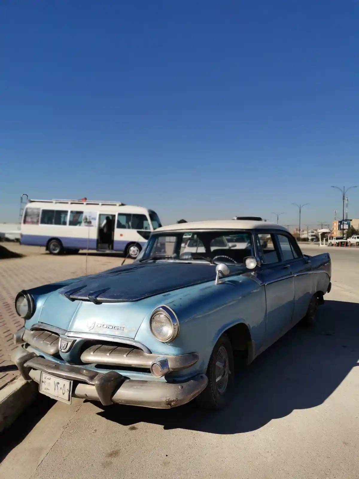 A vintage light blue Dodge sedan with a white roof is parked on a paved lot under a clear blue sky, showing signs of wear…
