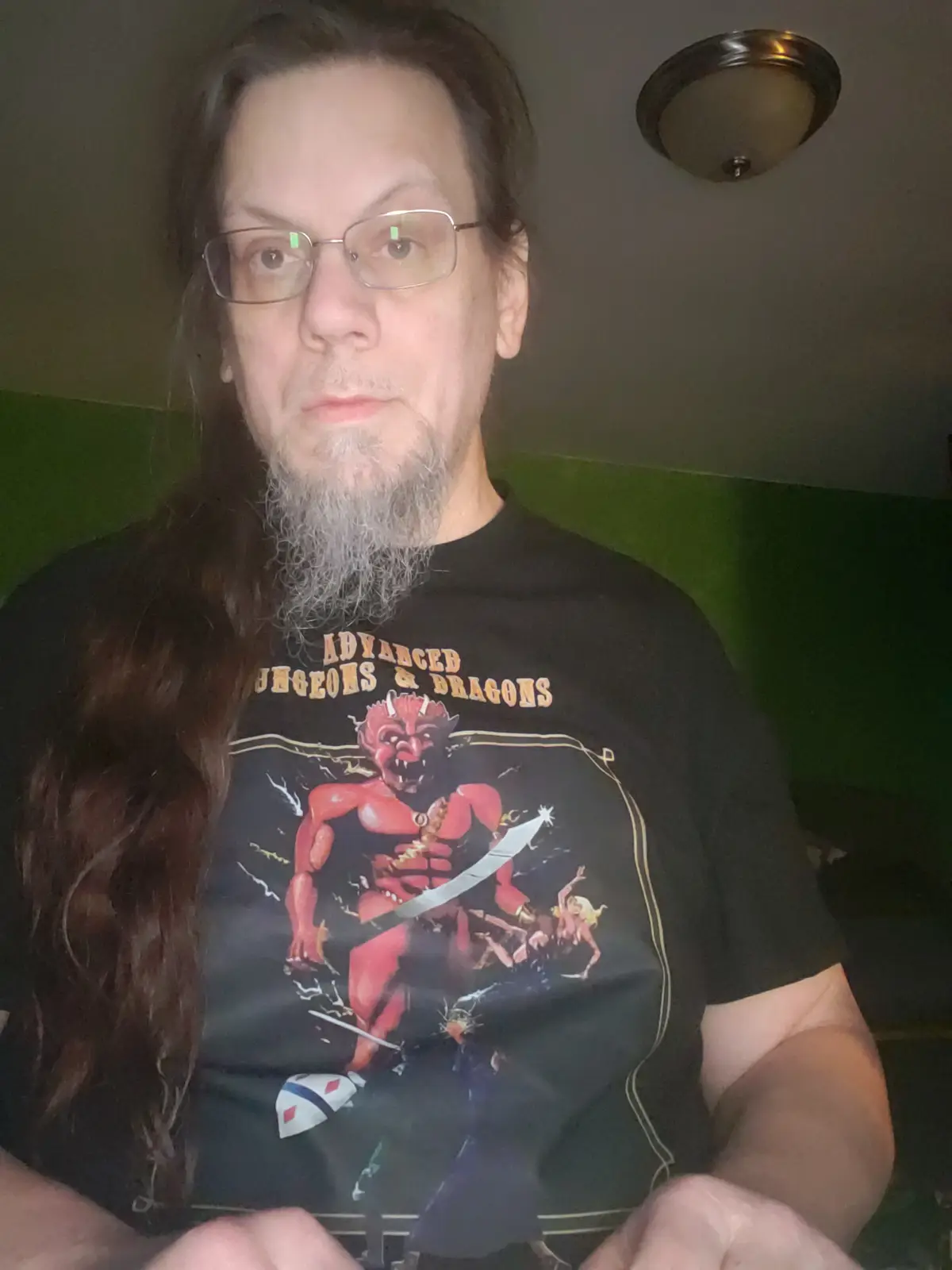A man with long brown hair, glasses, and a gray beard wears a black 'Advanced Dungeons & Dragons' t-shirt featuring a red…
