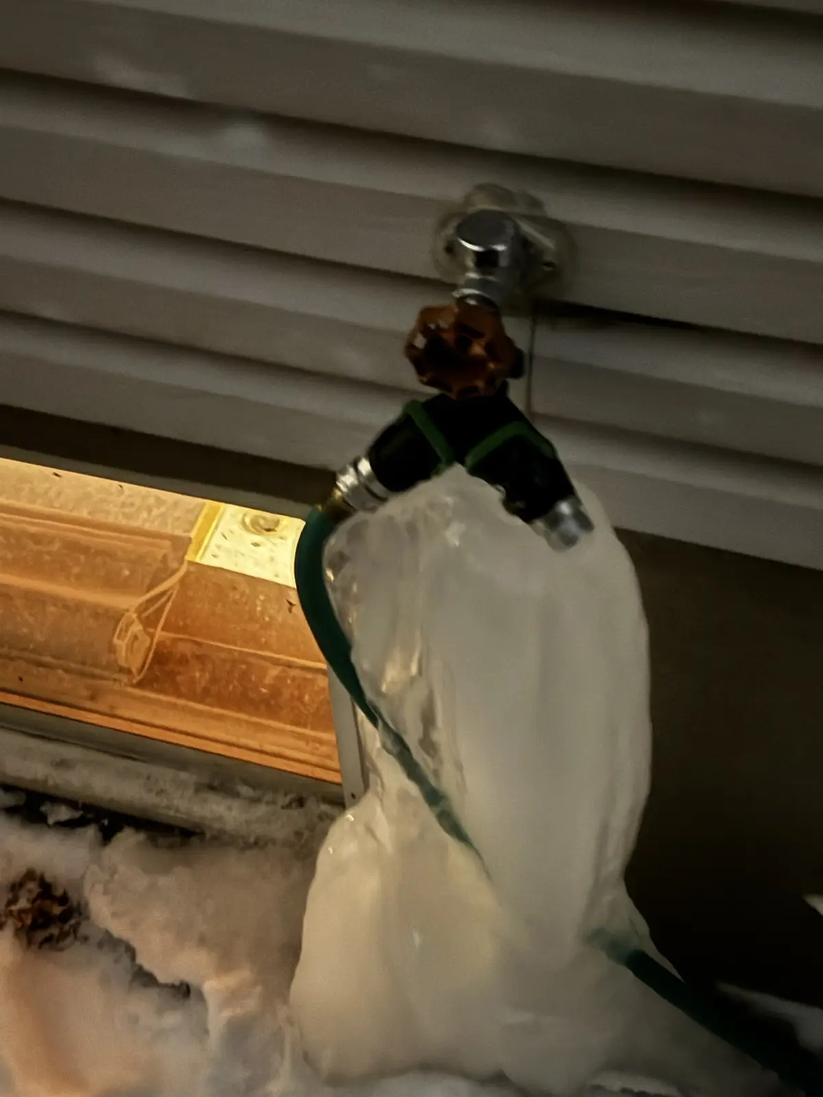A close-up photo shows a garden hose spigot with a large, frozen ice formation clinging to the hose and nozzle, set against…