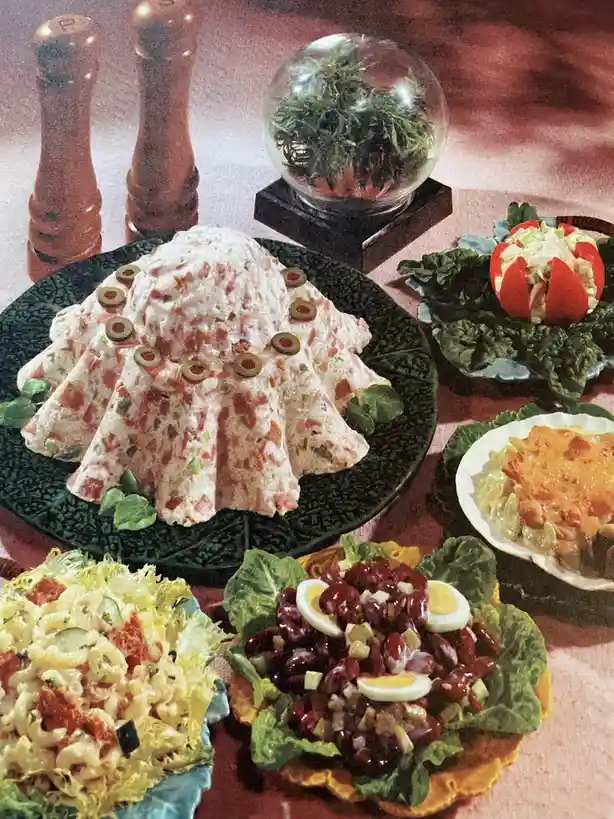 A vintage-style food photograph displays several elaborately arranged salads on decorative plates, including a large…