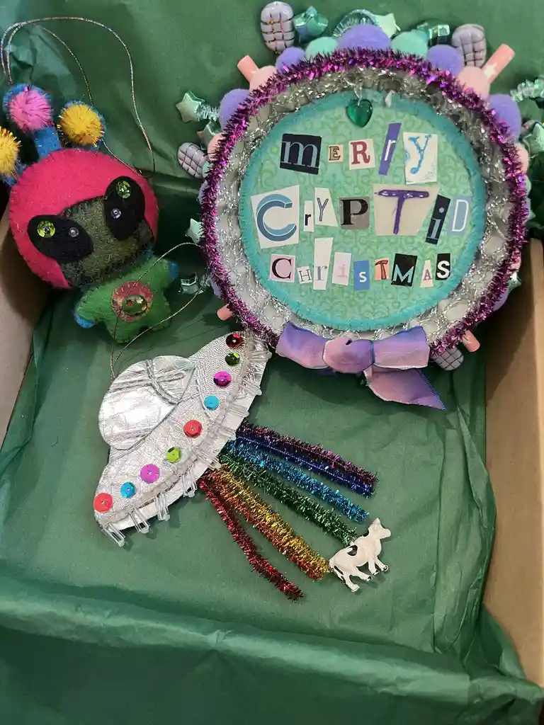 A collection of handmade, colorful Christmas ornaments including a felt cryptid creature, a glittery wreath with the text…