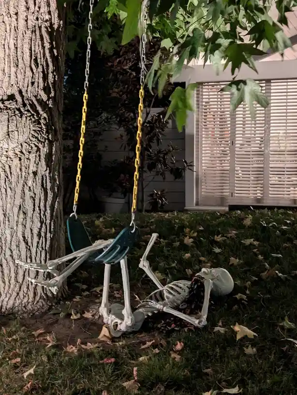 A plastic skeleton is partially on a green swing with yellow chains, hanging from a tree, while its upper body lies on the…