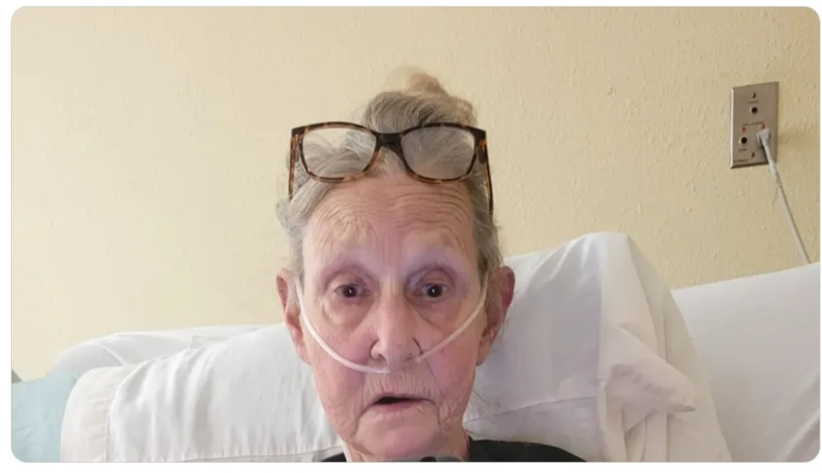 An elderly woman with glasses perched on her head and an oxygen tube across her nose sits in a hospital bed, looking…