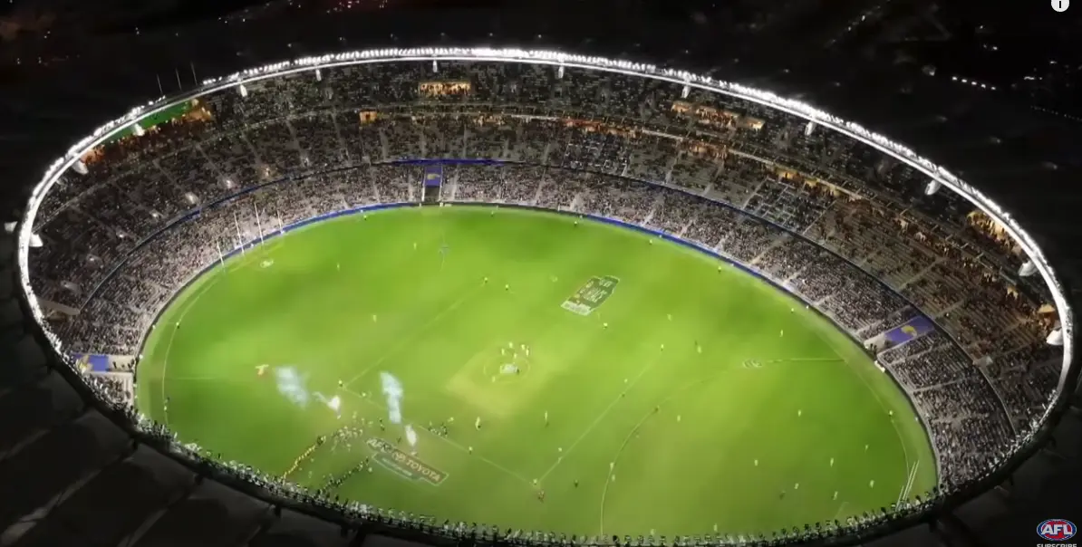 An aerial night view of a packed, brightly lit Australian Rules Football stadium with players on the green field. The…