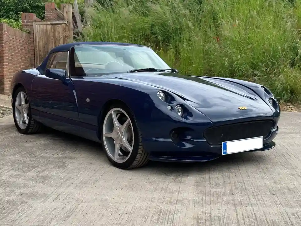 A dark blue convertible sports car, likely a TVR, is parked on a concrete driveway next to a brick wall and tall green…
