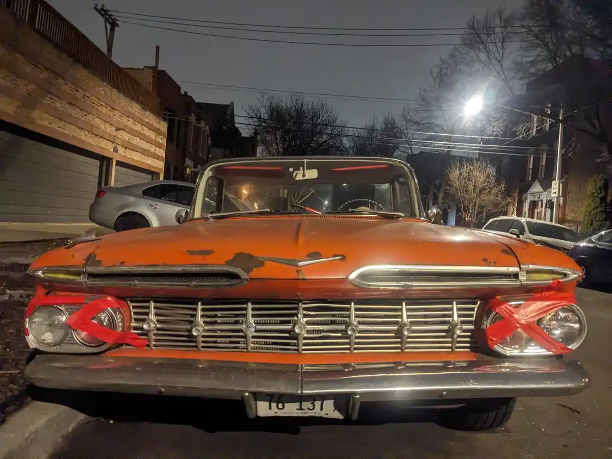 A rusty orange vintage car is parked on a city street at night, with red tape tied in an 'X' over both headlights…