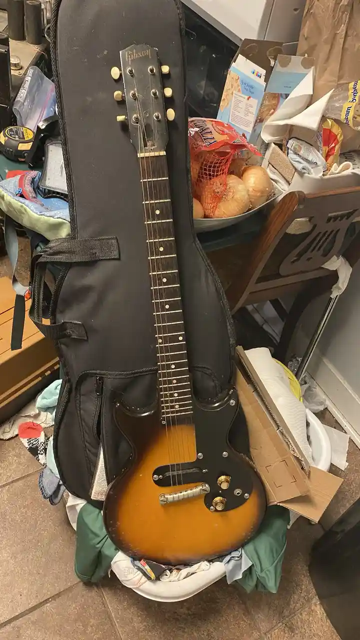A Gibson electric guitar with a sunburst finish is partially emerging from its black case, standing upright on a pile of…