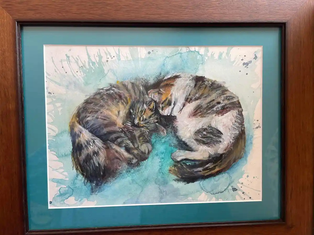 A watercolor painting of two calico cats curled up and sleeping together in a wooden frame with a teal mat. The cats are…