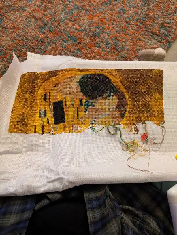 A partially completed cross-stitch piece depicting Gustav Klimt's 'The Kiss' is shown, with the bottom half of the artwork…