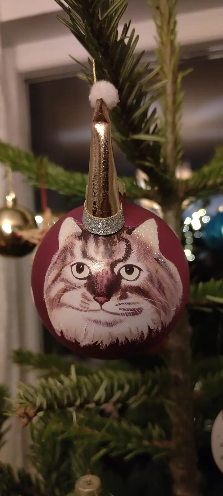 A close-up photo shows a maroon Christmas ornament hanging on a pine tree, featuring a painted portrait of a fluffy tabby…