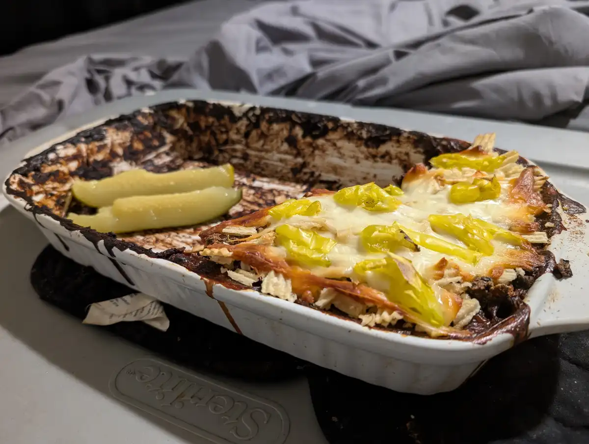 A white ceramic baking dish with burnt edges and mostly eaten lasagna, featuring melted cheese and green peppers, sits on a…