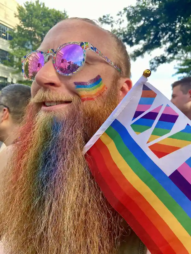 A man with a long, colorful beard and rainbow face paint smiles while holding a rainbow flag at an outdoor event. He wears…