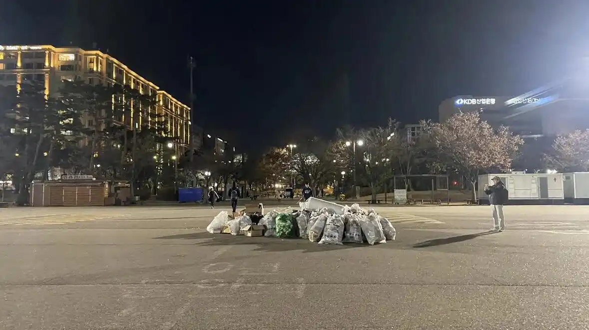 A nighttime photo shows a large pile of trash bags and cardboard boxes dumped in the middle of a paved public plaza. In the…