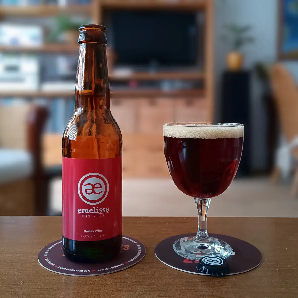 A brown bottle of Emelisse Barley Wine beer with a red label sits next to a stemmed glass filled with dark amber beer and a…